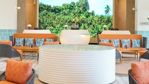 Un hall d’entrée doté d’un grand écran diffusant des images de paysages tropicaux, un comptoir de réception et des espaces de détente avec des coussins à motifs.