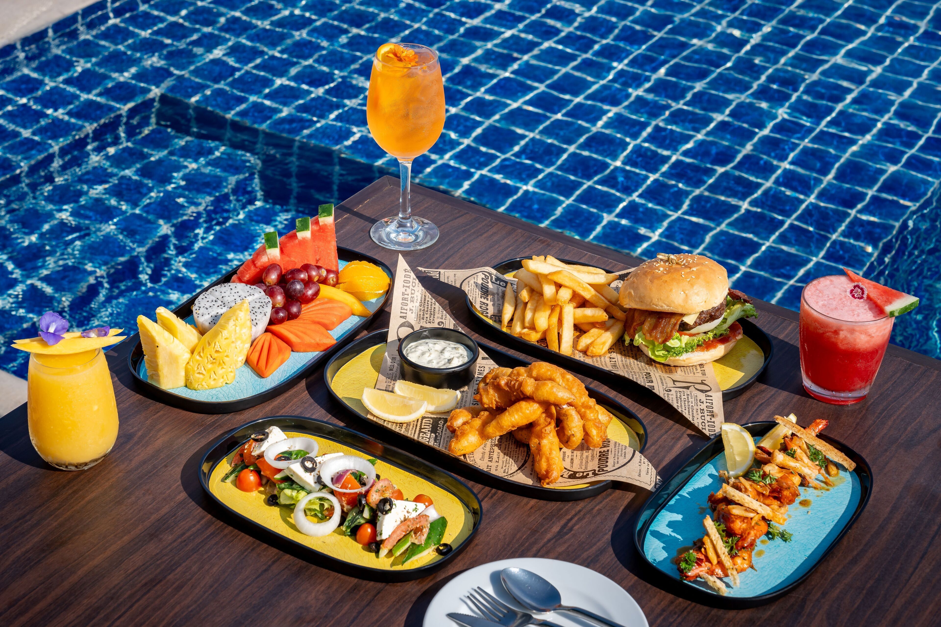 A table with various dishes including fish and chips, a burger, and a salad, set against a poolside backdrop.