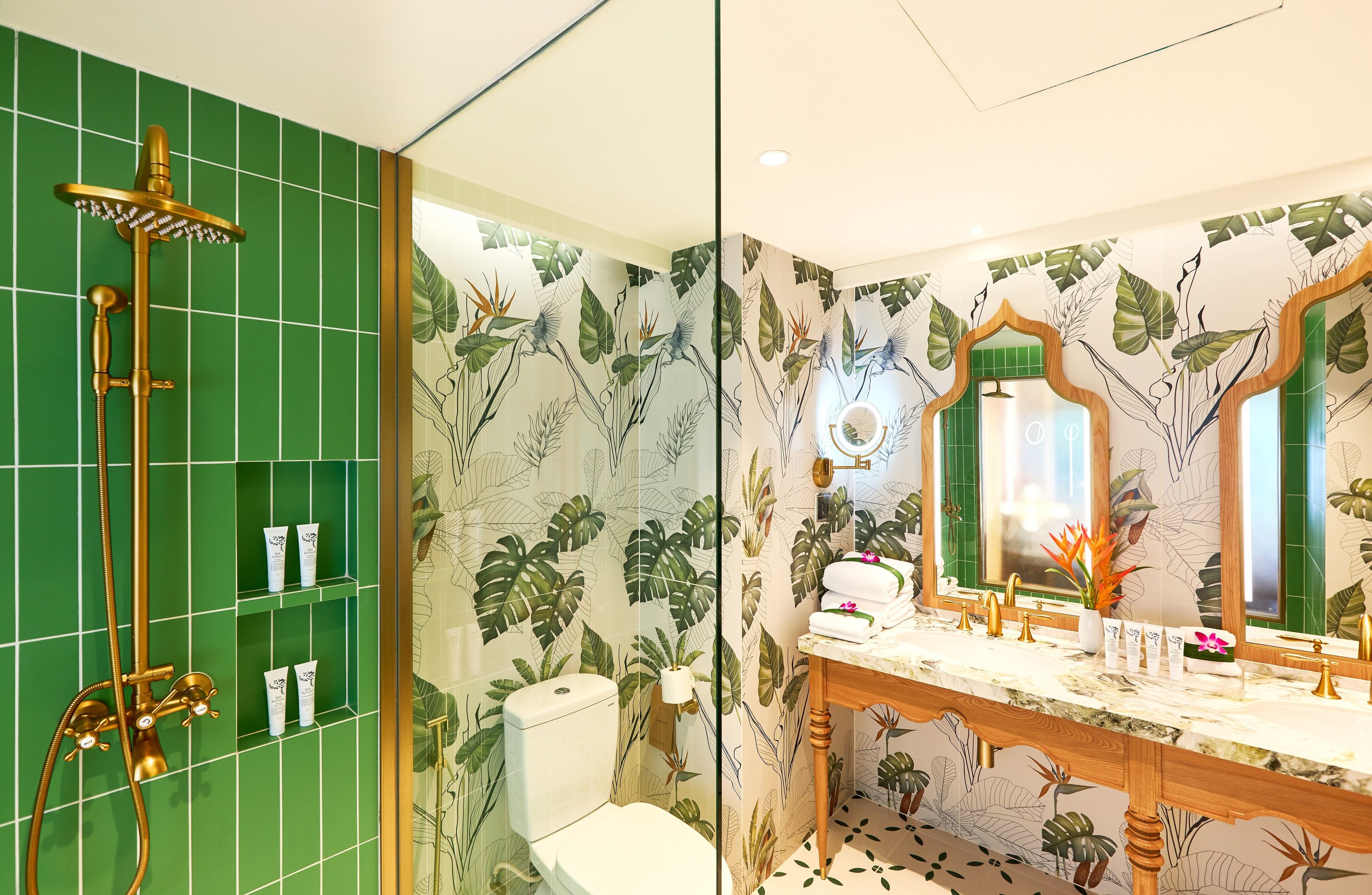 A bathroom with a green tiled wall, a gold showerhead, a toilet, a vanity with a mirror, and a sink.