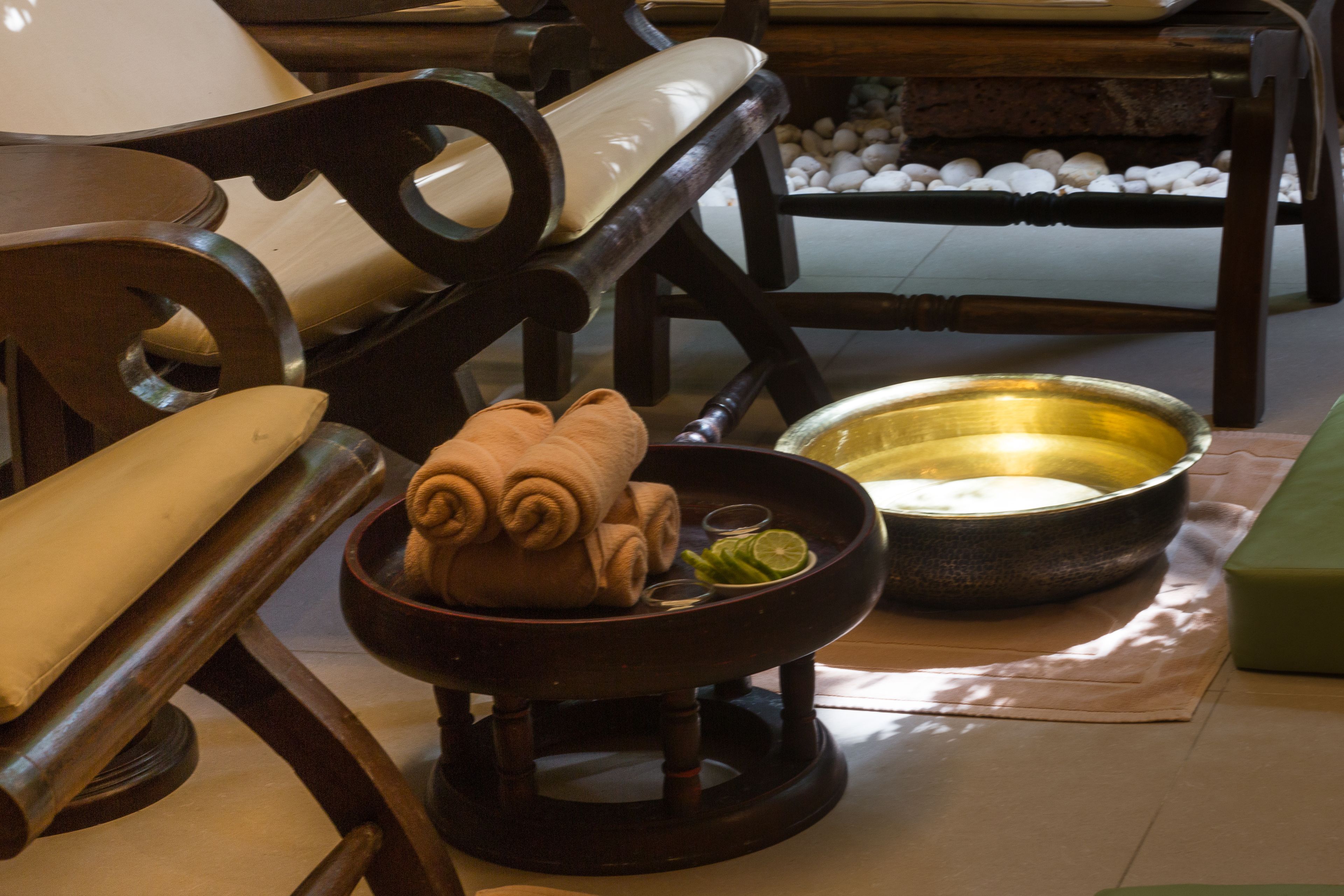 A wooden chair with a cushioned seat and backrest, a small wooden table with a bowl containing rolled towels and a lime wedge, and a stone basin with a golden interior.