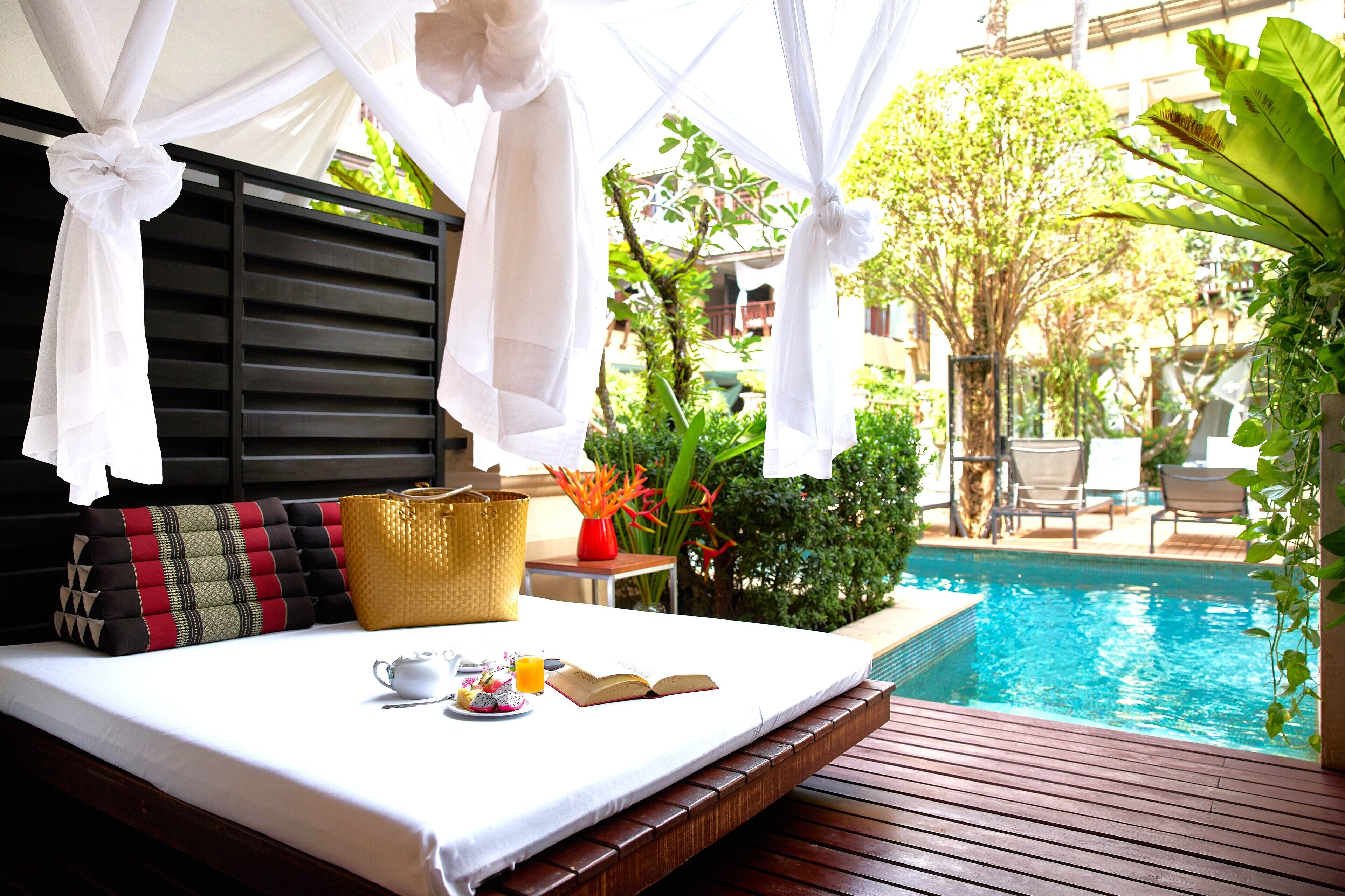 A bed with a white canopy, a bedside table with a basket and a book, and a view of a swimming pool with greenery in the background.