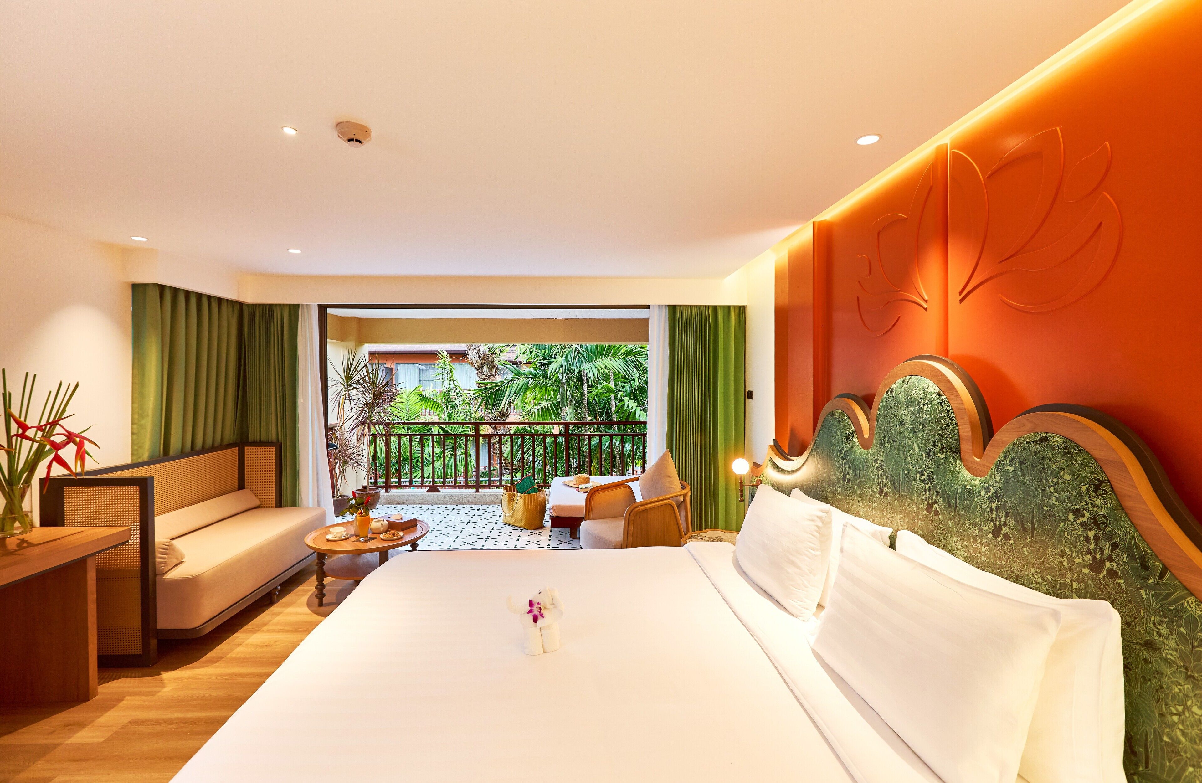 A hotel room with a large bed, a sofa, a small table with a vase, and a balcony with plants.