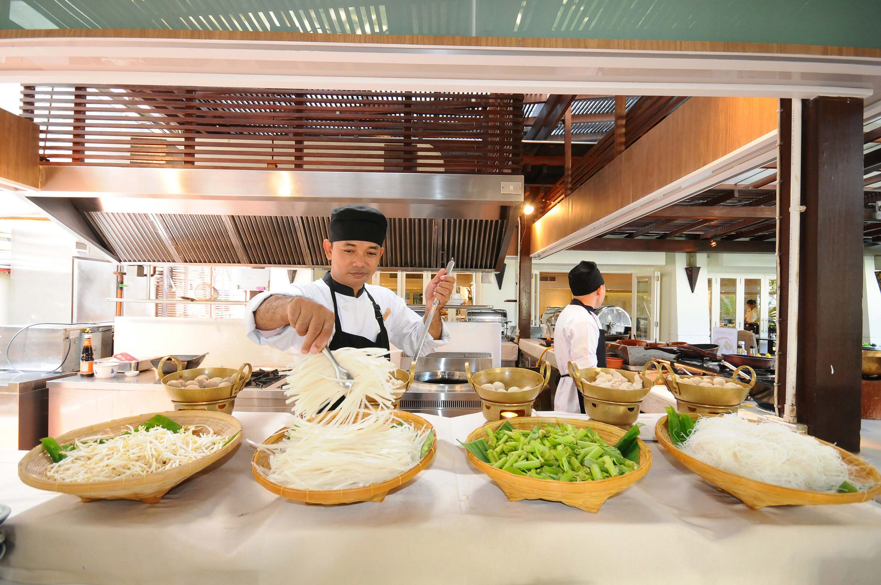 Uma cozinha com chefs preparando alimentos, diversos utensílios de cozinha e ingredientes expostos no balcão.