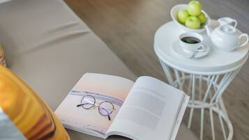 A book with glasses on top, a bowl of green apples, a cup of coffee, and a teapot on a small white table.