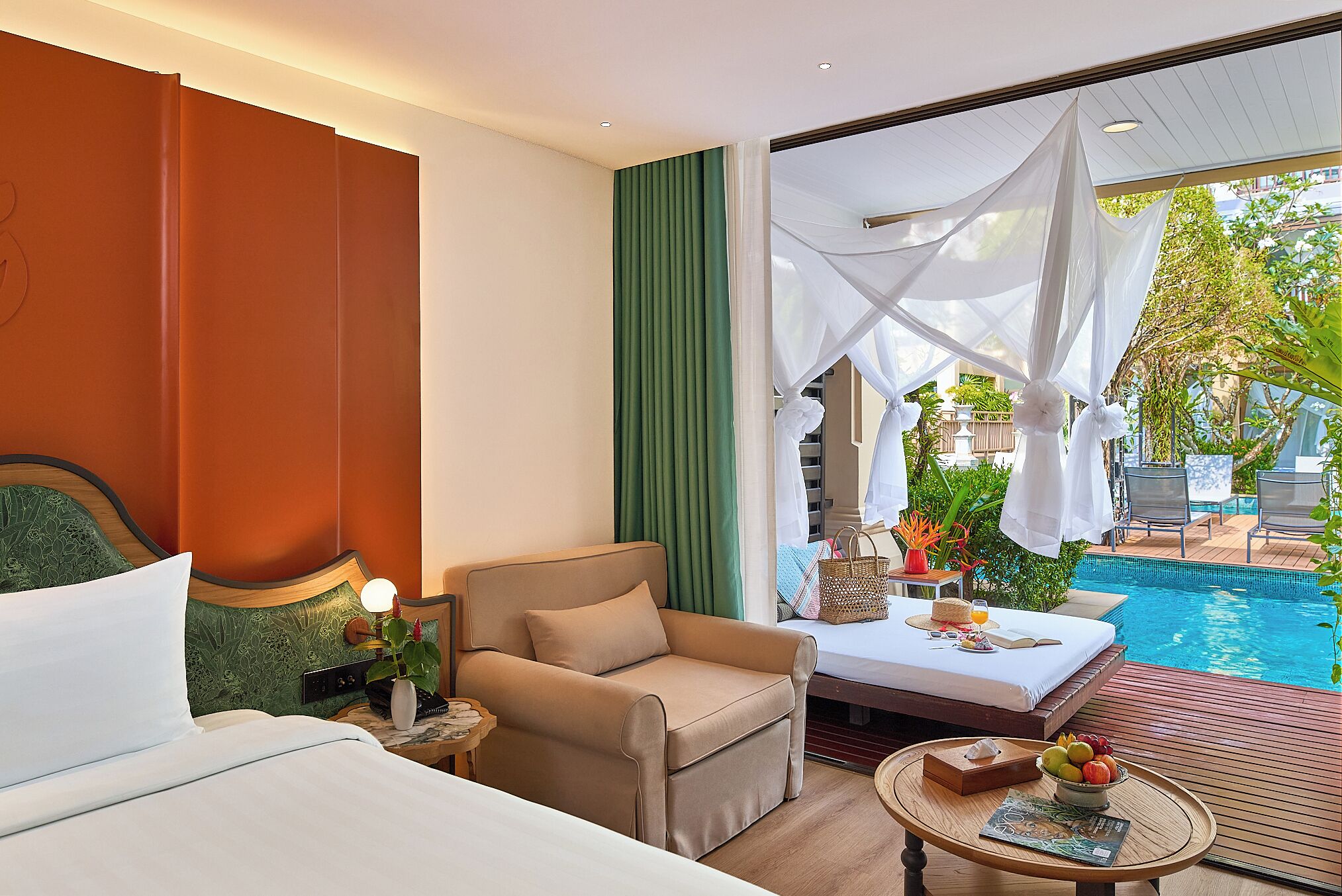 A hotel room with a bed, a sofa, a small table with fruit, and a view of an outdoor swimming pool.