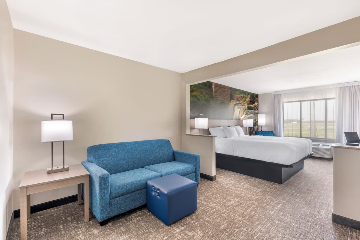 Ein Hotelzimmer mit einem blauen Sofa, einem kleinen Hocker, einem Schreibtisch mit Lampe, einem Bett mit weißer Bettwäsche und einem großen Fenster mit Vorhängen.