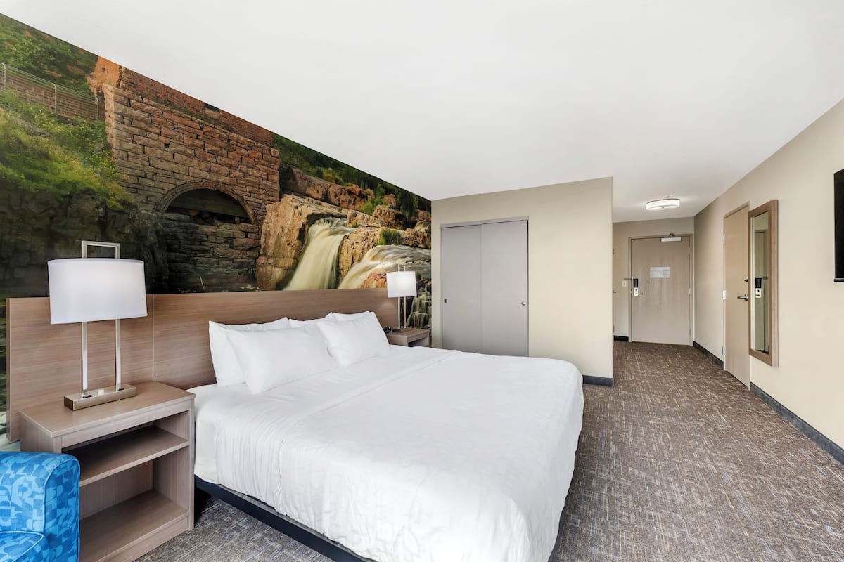 Ein Hotelzimmer mit einem großen Bett, einem Nachttisch mit Lampe, einem Wandbild eines Wasserfalls und einem Teppichboden.