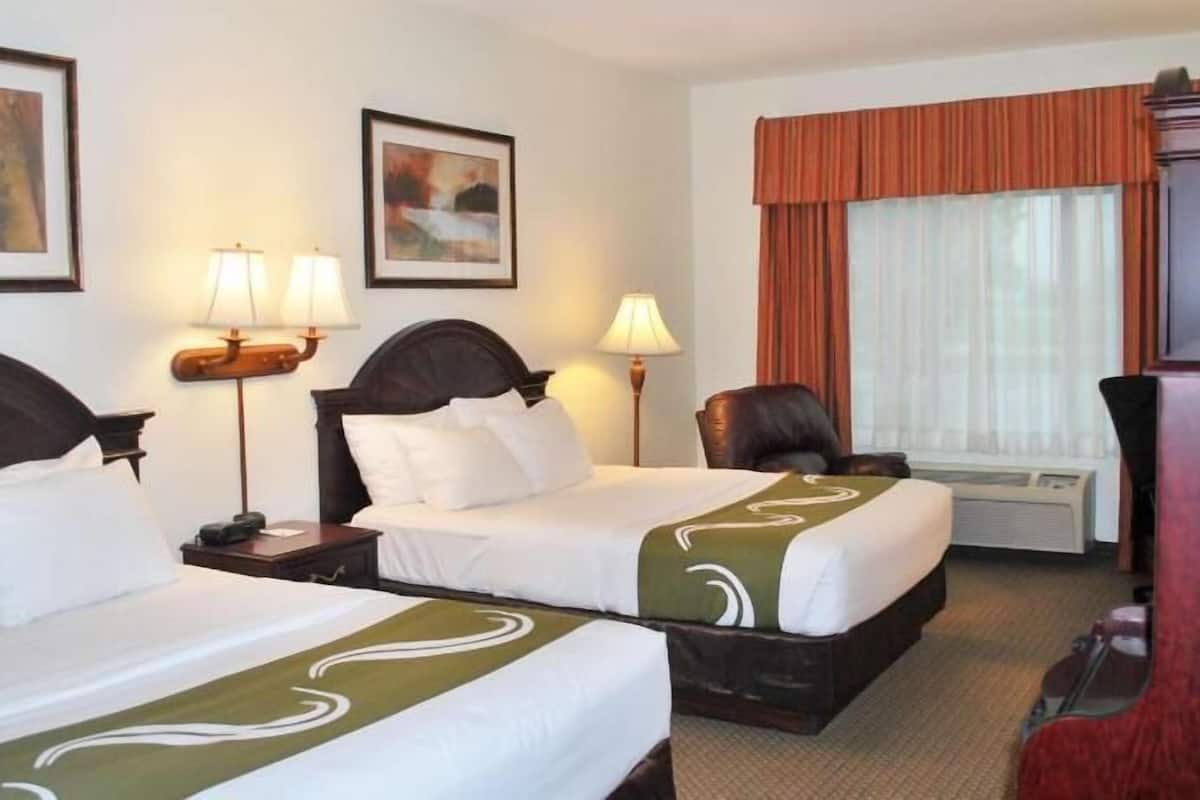 Ein Hotelzimmer mit zwei Betten, einem Schreibtisch, einem Stuhl, einer Schreibtischlampe, einem Fenster mit Vorhängen und Bildern an den Wänden.