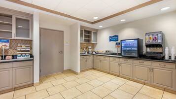 A clean, well-lit kitchen area with beige cabinets, a refrigerator, a microwave, and a coffee machine. There are signs indicating a healthy menu and a starting point for breakfast.