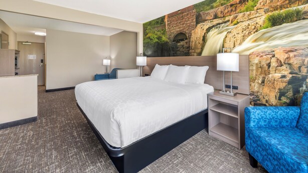 A hotel room with a large bed, a blue sofa, a nightstand with a lamp, and a mural of a waterfall on the wall. - Clarion Pointe Sioux Falls Airport (Sioux Falls)