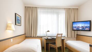 A hotel room with two single beds, a desk with a chair, a TV on the wall, and a window with curtains.