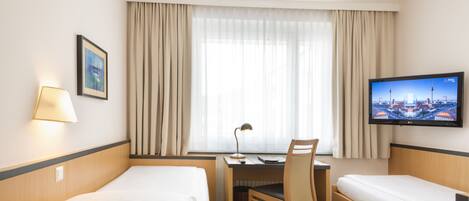 A hotel room with two single beds, a desk with a chair, a TV on the wall, and a window with curtains.