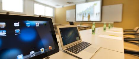Egy konferenciaterem, amelyben van egy laptop, egy táblagép és egy projektorvászon, amelyen egy prezentáció látható.