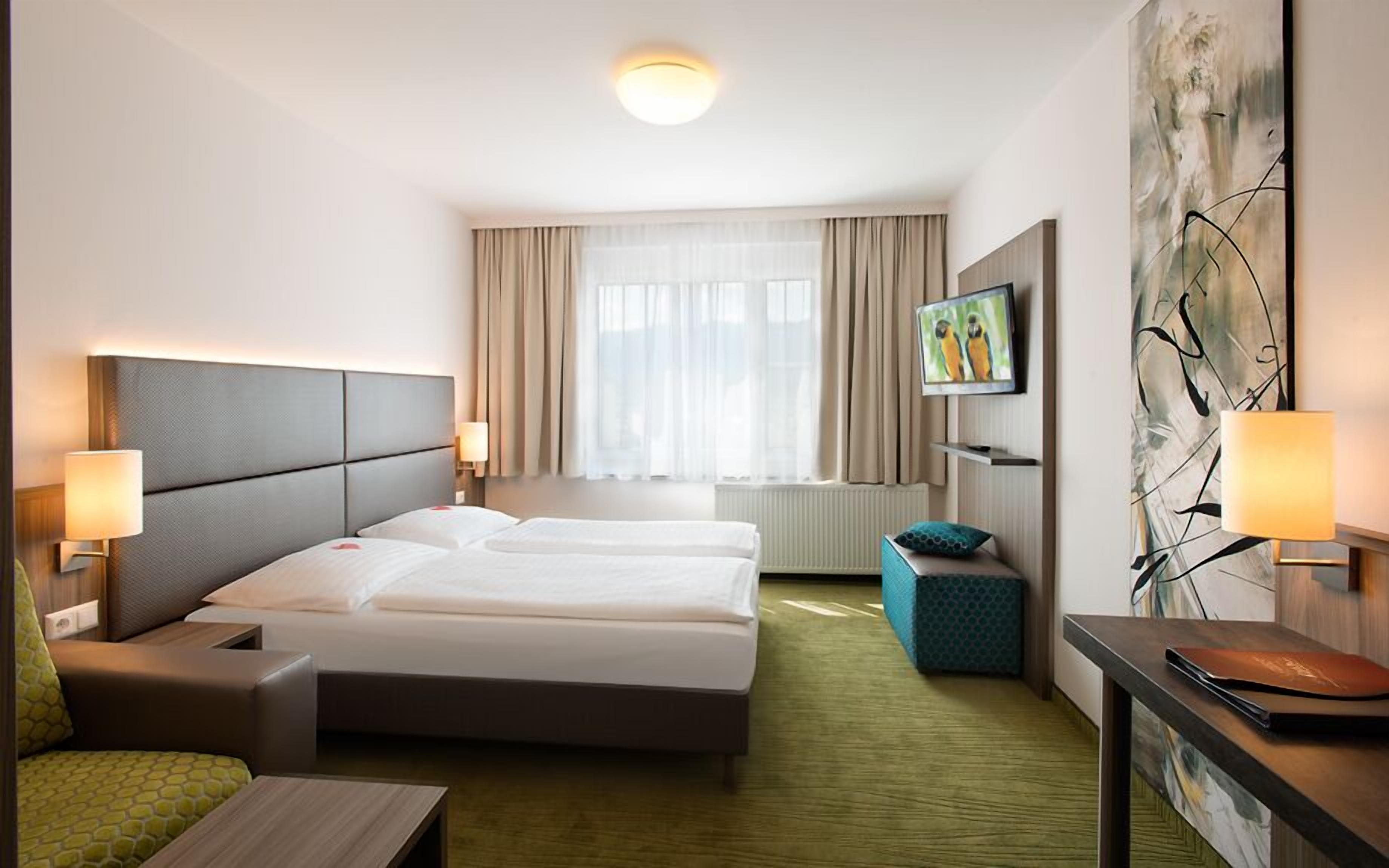 Een hotelkamer met een bed, twee nachtkastjes, een televisie, een schilderij en een raam met gordijnen.