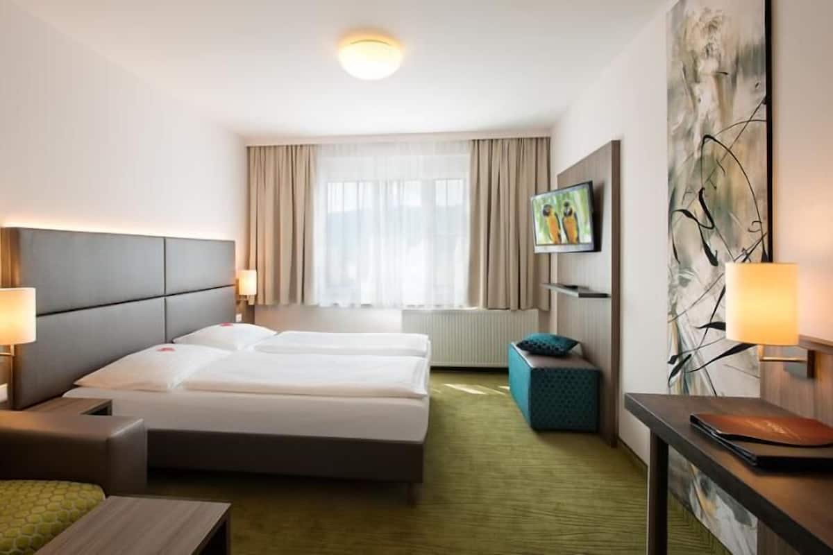 Une chambre d’hôtel avec un lit, deux tables de chevet, une télévision, un tableau et une fenêtre avec des rideaux.