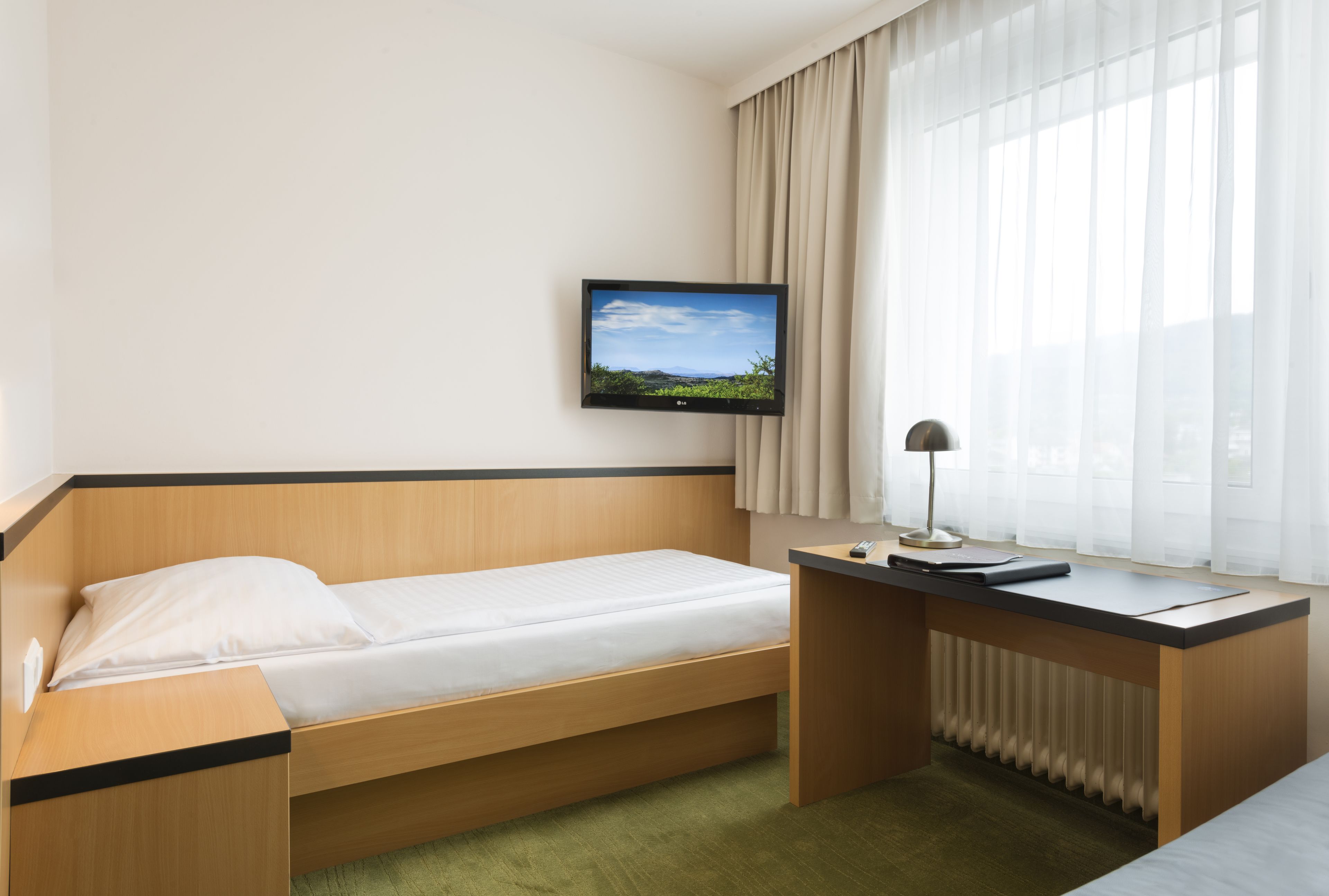 A hotel room with a single bed, a desk, a television mounted on the wall, and a window with curtains.