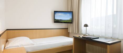A hotel room with a single bed, a desk, a television mounted on the wall, and a window with curtains.