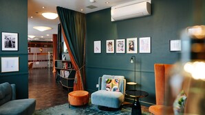 A modern hotel lobby with a teal wall, a bookshelf, a grey armchair, and a round ottoman. - Hotel City Villach (Villach)