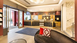 A modern hotel lobby with a reception desk, a sofa with cushions, a kitchen area, and a staircase. - Hotel City Villach (Villach)