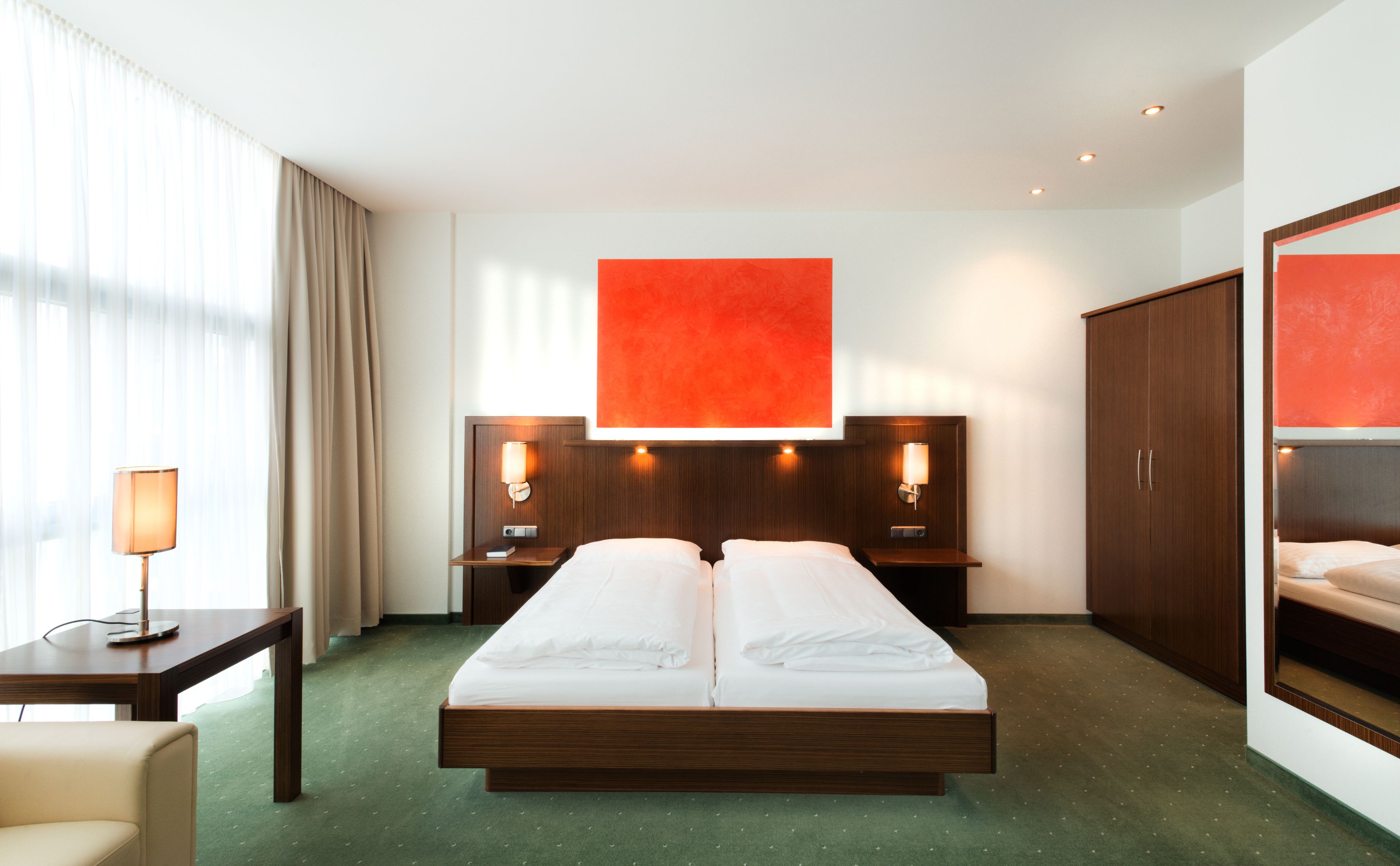 Una camera d'albergo con un letto grande, due comodini, una scrivania con lampada, un armadio e un'ampia finestra con tende.
