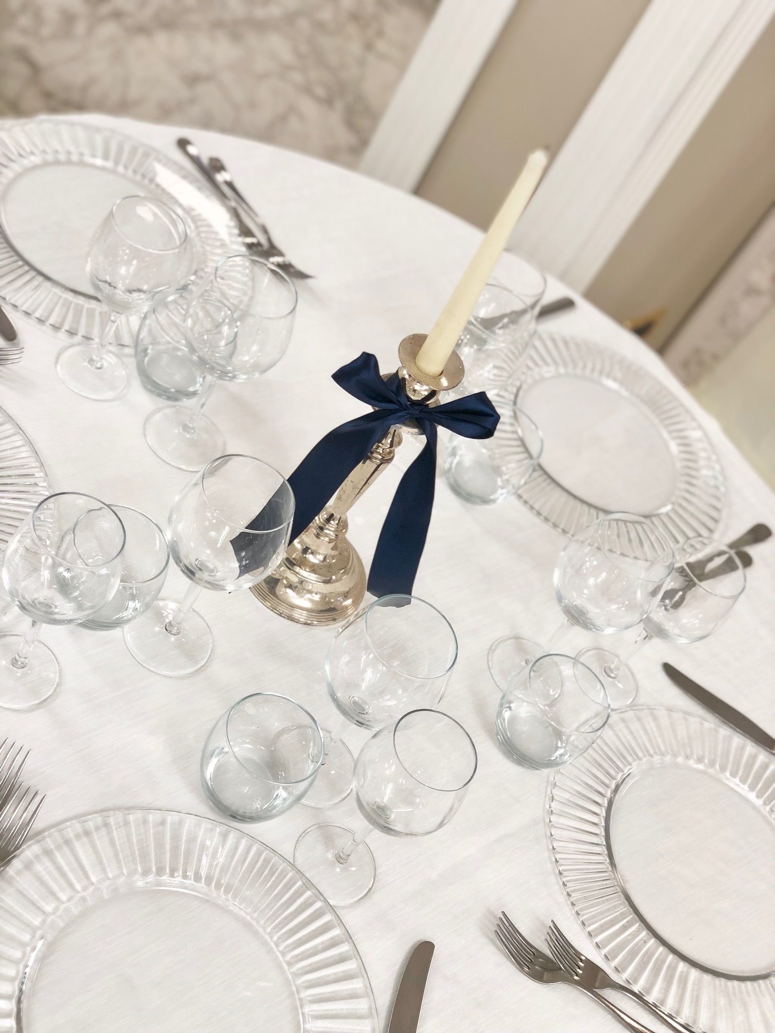 Tavola imbandita per un evento formale, con bicchieri trasparenti, piatti e posate, con una candela decorata da un nastro blu al centro.