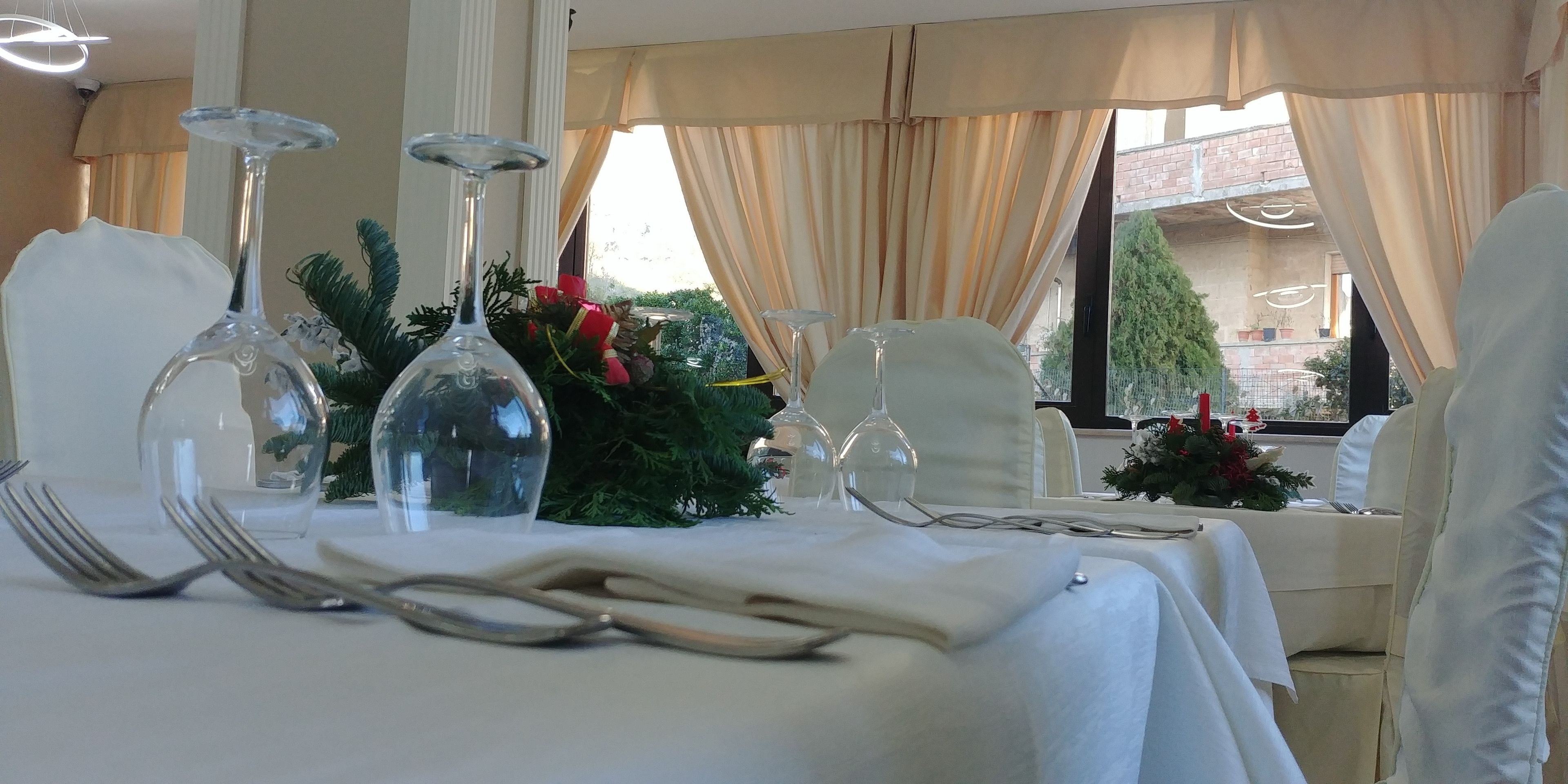 A dining table set with white tablecloths, clear glassware, and a centerpiece of greenery and red flowers.
