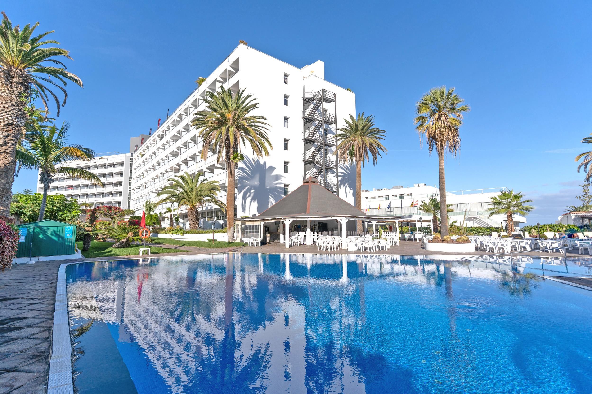 Un hotel con una piscina grande, palme e un edificio bianco con numerosi balconi.