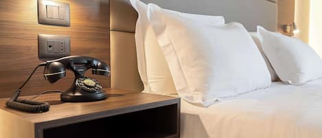 A neatly made bed with white linens, a wooden bedside table with a rotary phone, and wall-mounted light switches.