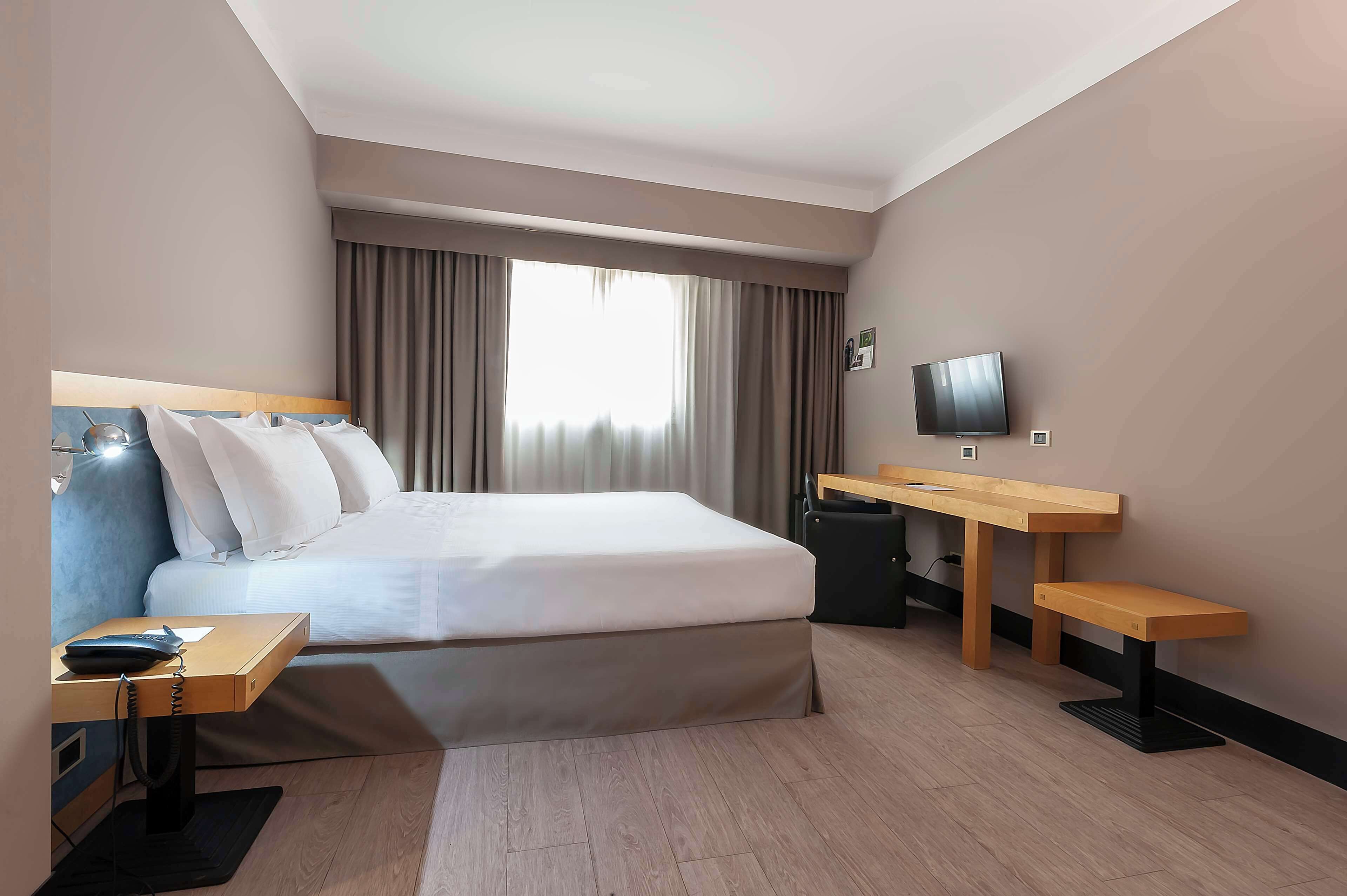 Habitación de hotel con una cama grande, un escritorio, una silla, un televisor y una ventana con cortinas.