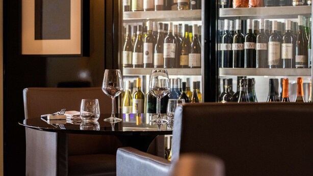 L’intérieur d’un restaurant présente une table dressée pour deux personnes, un réfrigérateur à vin rempli de différentes bouteilles et un tableau encadré accroché au mur.