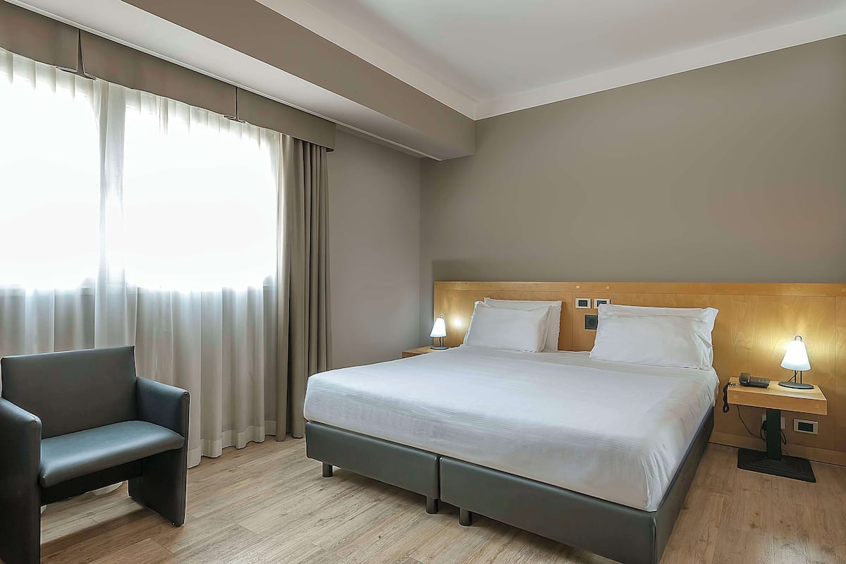Une chambre d’hôtel avec un lit, des lampes de chevet, une chaise et une fenêtre avec des rideaux.