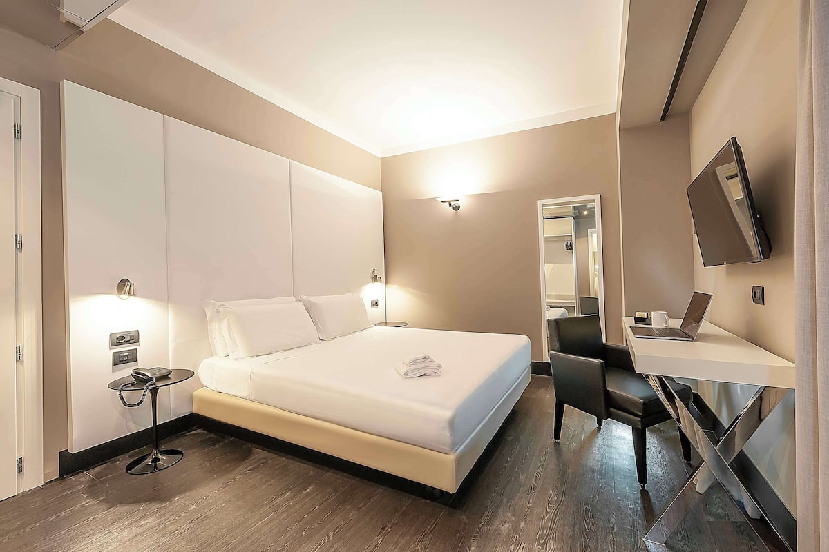 Une chambre d’hôtel moderne avec un grand lit, une table de chevet avec un téléphone, un bureau avec un ordinateur portable et un miroir.