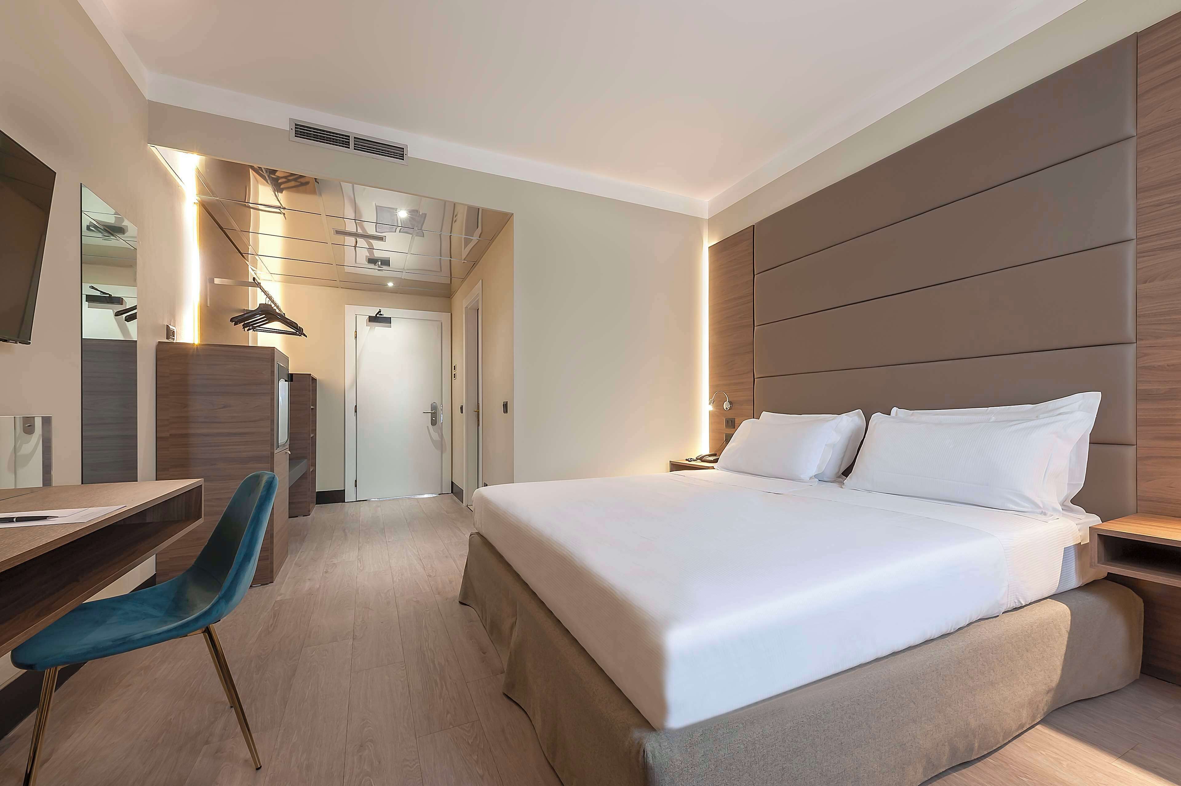 Habitación de hotel moderna con una cama grande, un escritorio y una silla. Hay un espejo en la pared y una puerta que da a otra habitación.