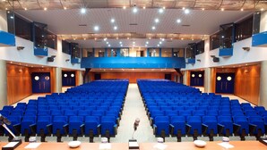 Une salle de conférence avec des rangées de chaises bleues, un pupitre muni d’un microphone et une table en bois sur laquelle se trouvent des papiers et un verre.