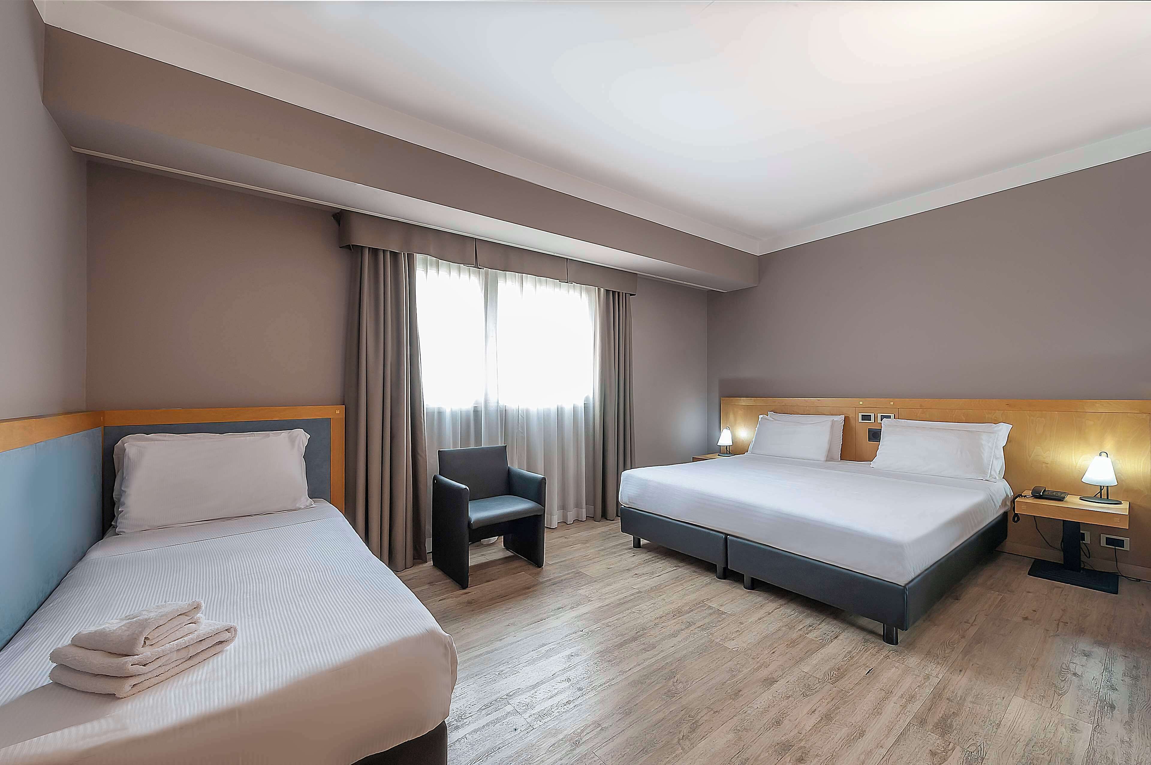 A hotel room with two beds, a chair, a nightstand with a lamp, and a window with curtains.