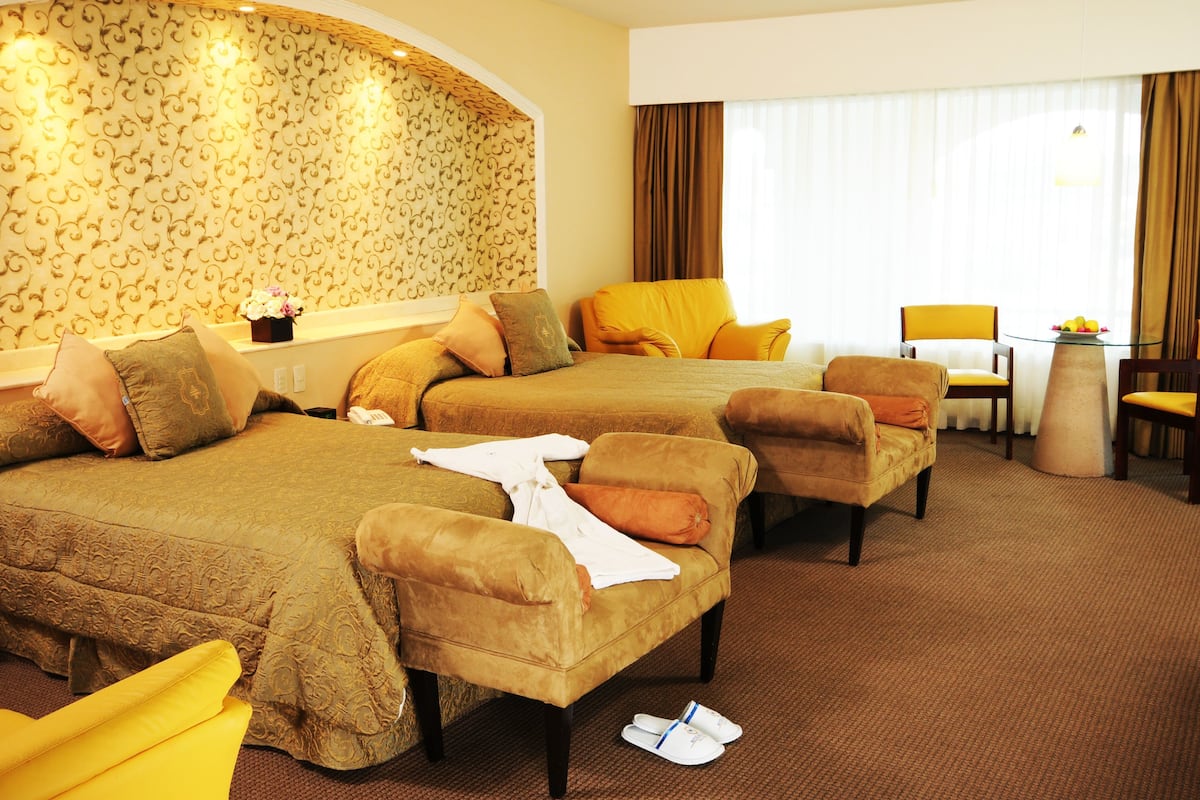 Habitación de hotel con dos camas, un área de descanso con una mesa y sillas, y un gran ventanal con cortinas.