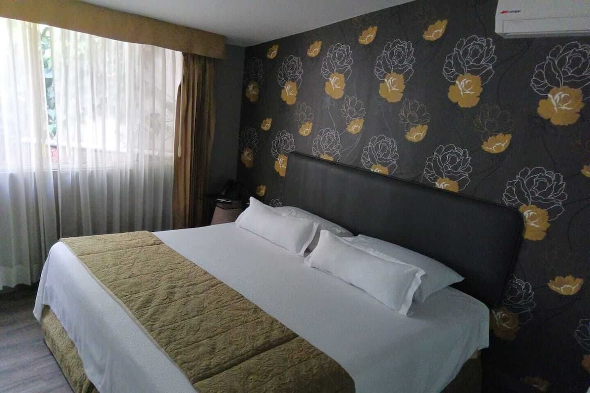 Una habitación de hotel con una cama grande, un papel tapiz floral oscuro y una ventana con cortinas de gasa.