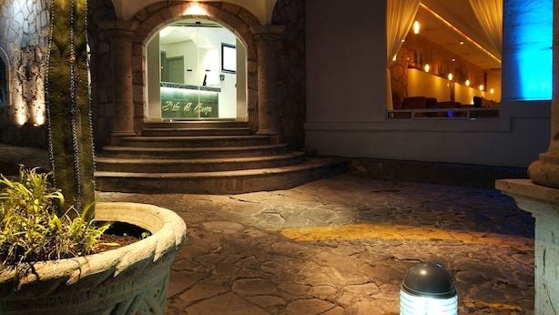 Un vestíbulo con suelo de piedra, una planta grande en maceta, una lámpara de pie encendida y vistas a una habitación con cortinas.