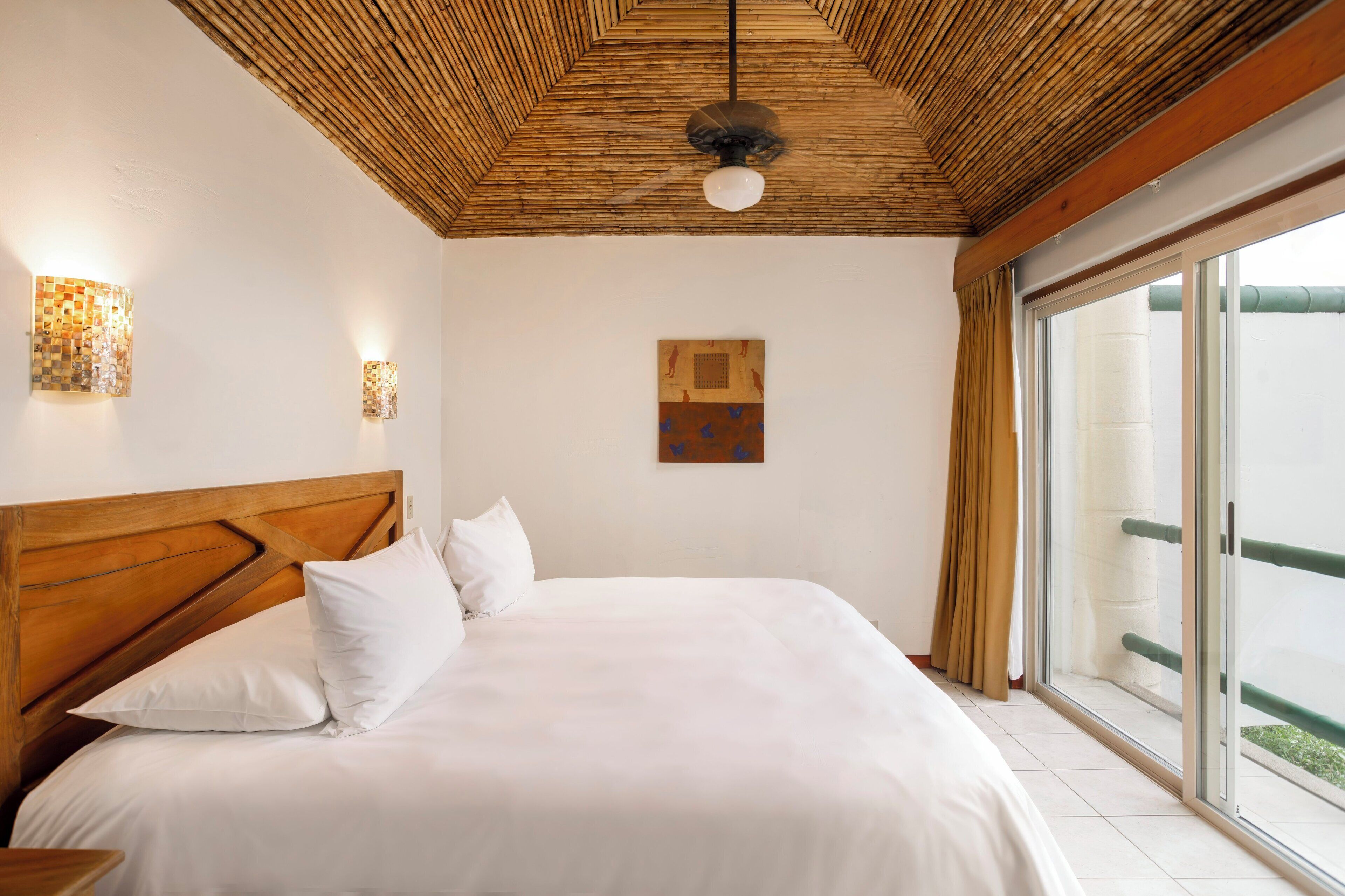 A bedroom with a wooden headboard, a bed with white linens, a ceiling fan, wall-mounted lights, and a large window with a view of greenery.
