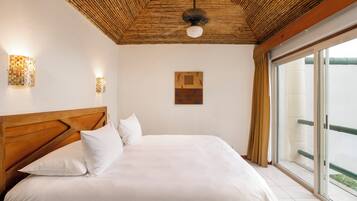 A bedroom with a wooden headboard, a bed with white linens, a ceiling fan, wall-mounted lights, and a large window with a view of greenery.