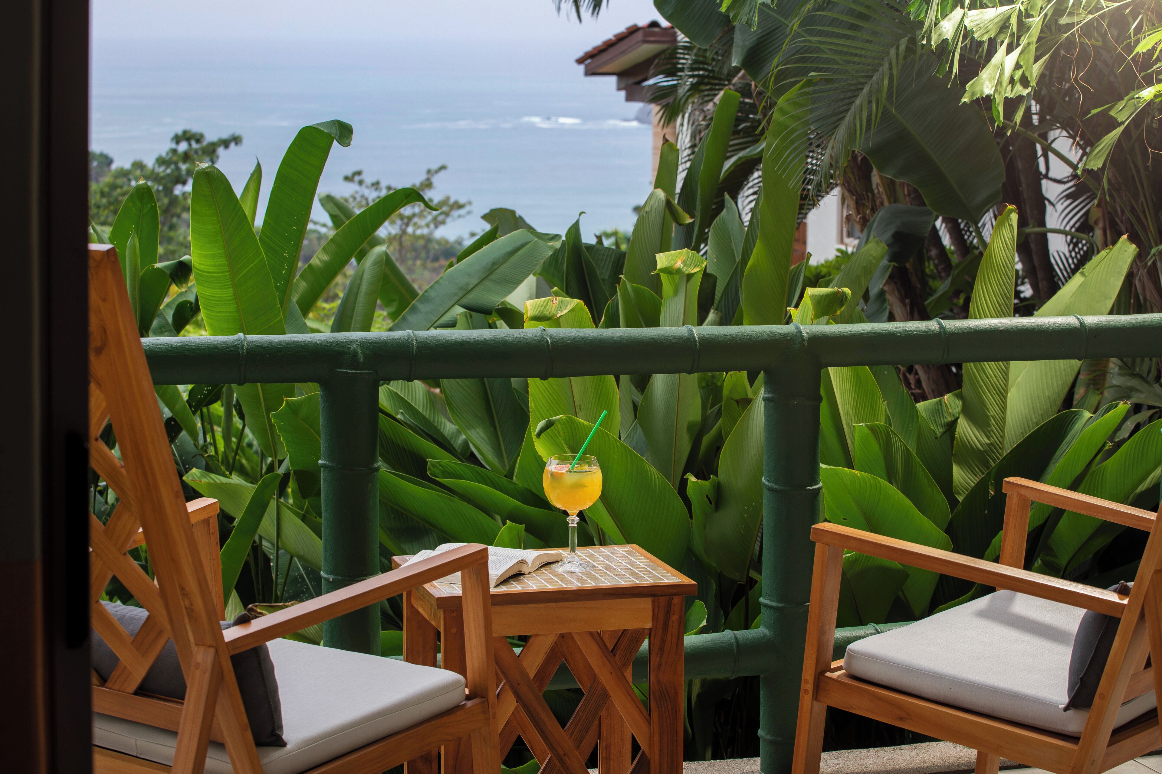 A wooden chair and table with a drink on a balcony overlooking the ocean.