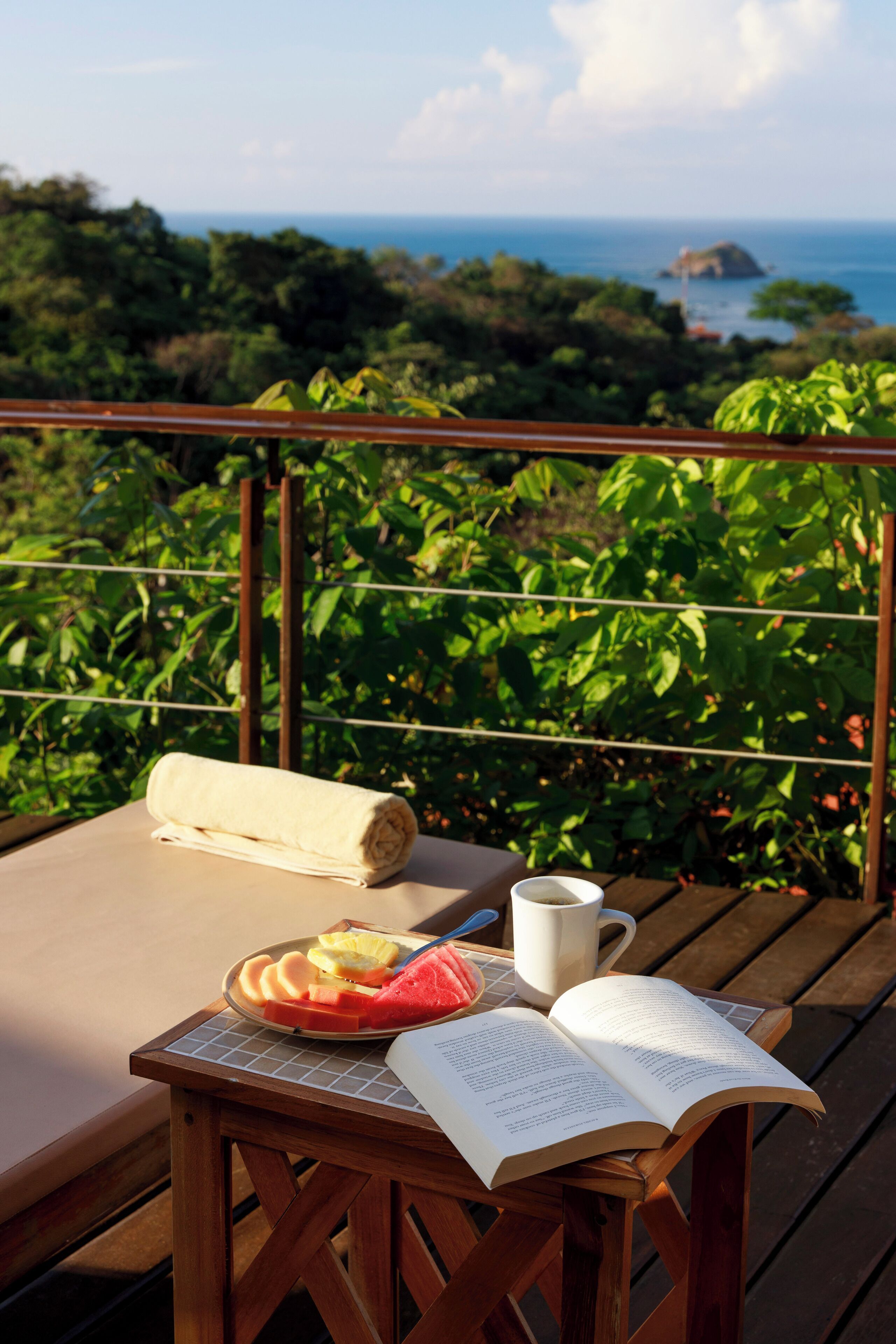 Eine Holzterrasse mit Tisch, gedeckt für eine Mahlzeit, einem Buch und einer Tasse, mit Blick auf eine üppige Landschaft und das Meer.