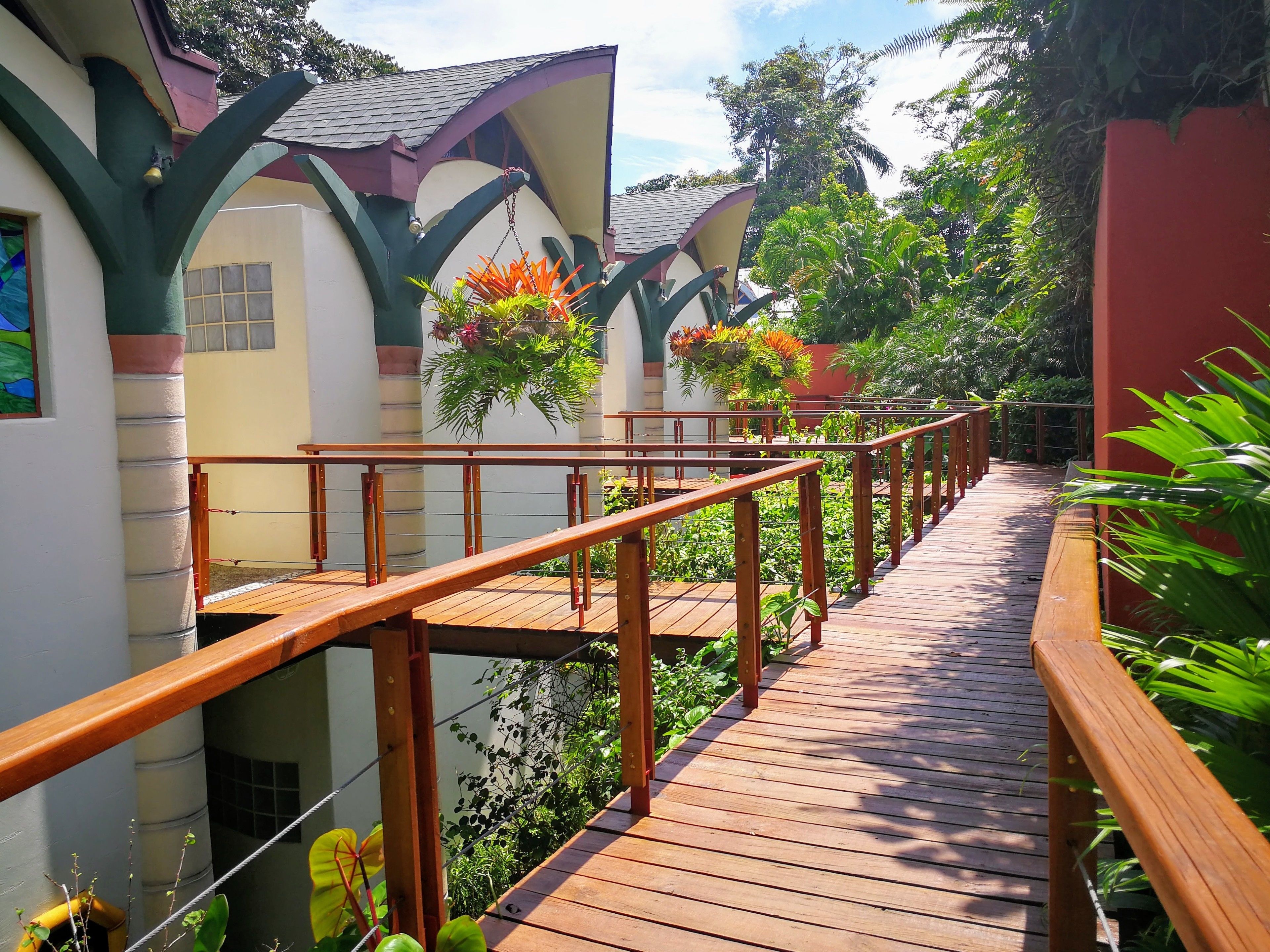A wooden deck with a metal railing, surrounded by lush greenery and a building with a sloped roof.