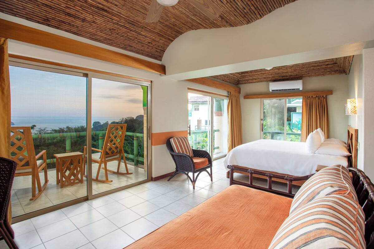 Une chambre d’hôtel avec un grand lit, un balcon avec des chaises et une vue sur la mer.