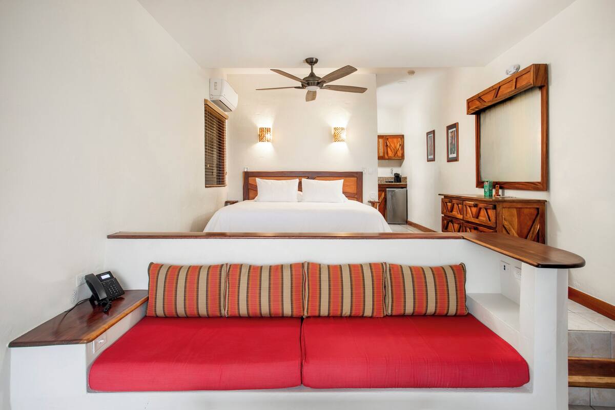 Une chambre d’hôtel avec un lit, un banc doté de coussins rouges, un ventilateur de plafond et une armoire en bois.