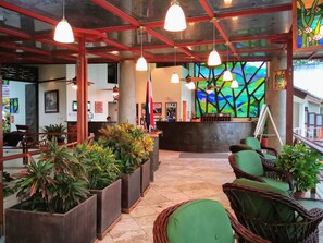 A lobby with a reception desk, green cushioned seating, potted plants, and hanging pendant lights.
