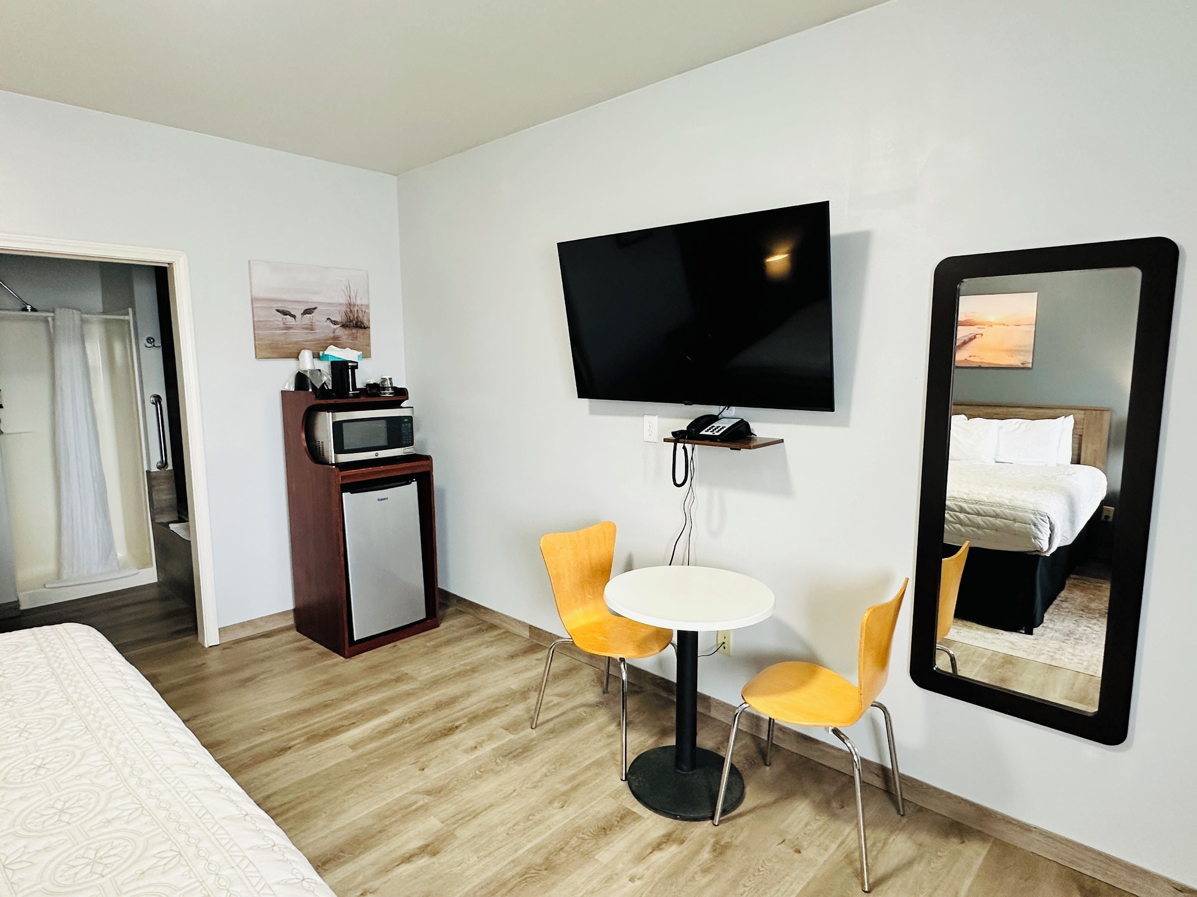 A hotel room with a bed, a TV mounted on the wall, a small desk with a microwave, and a table with two chairs.