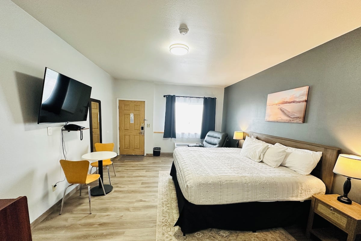 A hotel room with a bed, a desk with a chair, a TV, a window with curtains, and a painting on the wall.