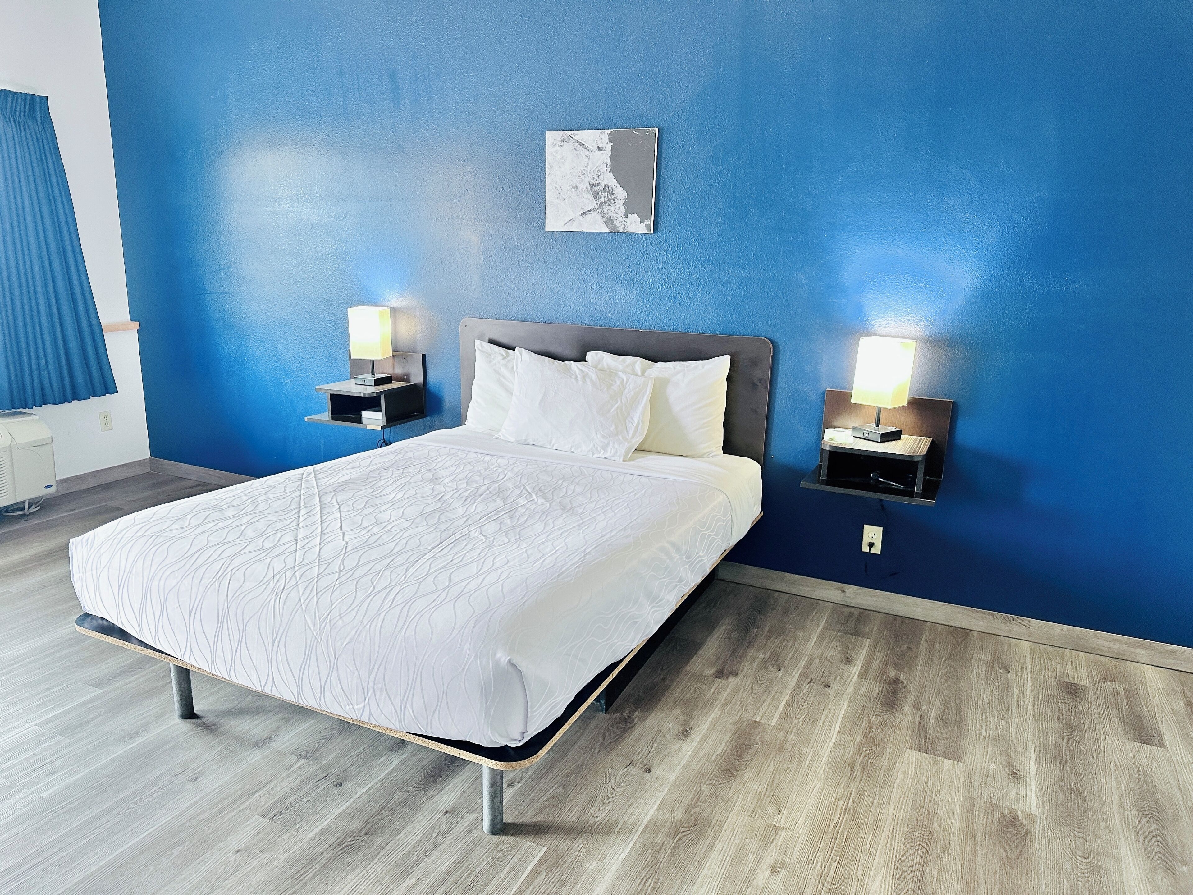 A hotel room with a large bed, two bedside tables with lamps, a blue accent wall, and a small artwork above the bed.