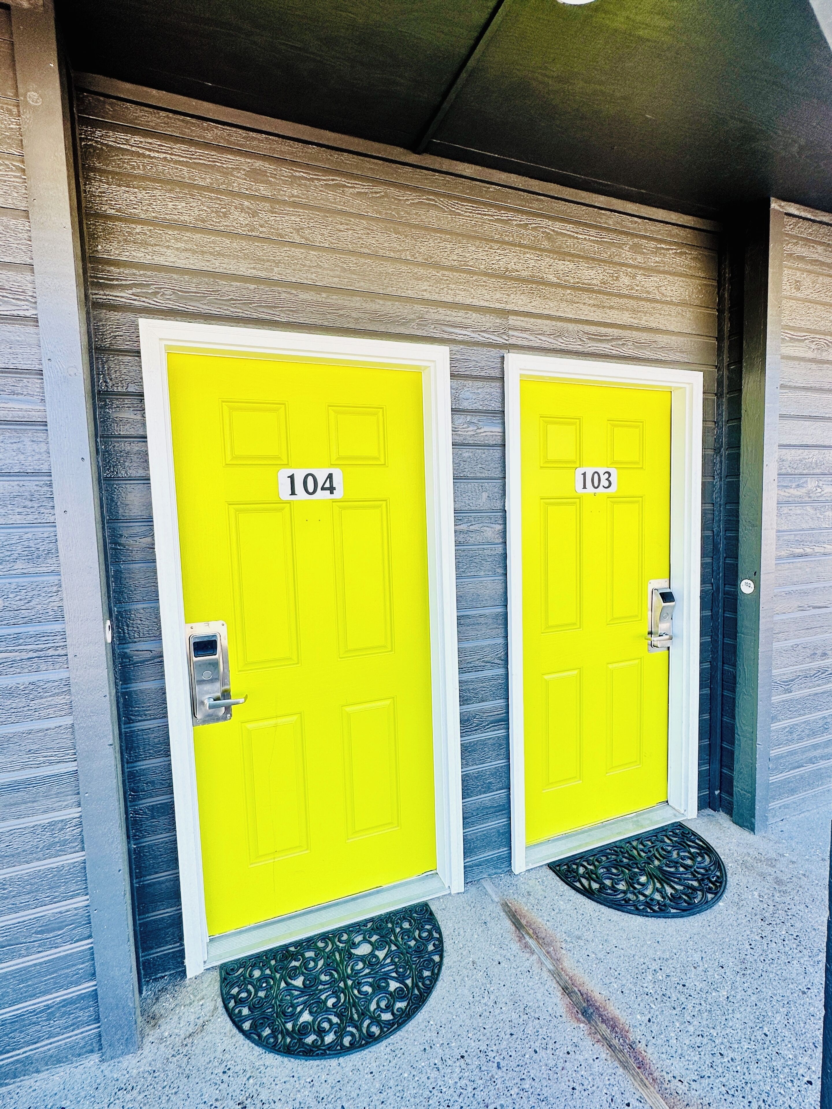 Deux portes jaune vif, portant les numéros 104 et 103, une sonnette métallique et un paillasson.